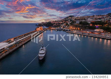 Aerial twilight view shows Funchal harbour in Madeira, Portugal, with a yacht by the cruise pier and promenade, hills of hotels and homes, and Sao Tiago Fortress at the inlet. 134284951