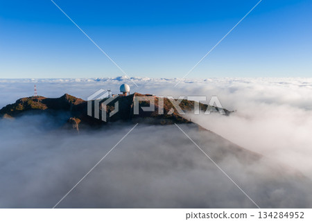 Aerial view shows Pico do Arieiro on Madeira, Portugal, with a white radar dome and antennas above clouds, reddish rock, and wide framing in clear morning light. 134284952