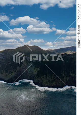 Aerial view of rugged sea cliffs and white surf on Madeira Island, Portugal, dark volcanic shore, green slopes, and high peaks under cumulus clouds in bright daylight. Aerial view of rugged sea cliffs and white surf on Madeira Island, Portugal, dark volcanic shore, green slopes, and high peaks under cumulus clouds in bright daylight. 134284967