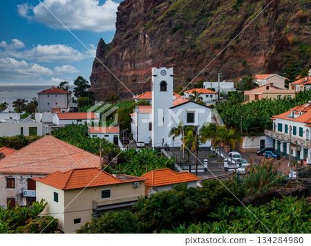 A whitewashed church with clock tower anchors terracotta roofs, banana terraces, palms, and pastel houses below a rust cliff, Atlantic at left, mild daylight scene. A whitewashed church with clock tower anchors terracotta roofs, banana terraces, palms, and pastel houses below a rust cliff, Atlantic at left, mild daylight scene. 134284980