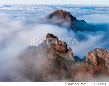 Aerial view shows rugged reddish peaks and ridgelines near Pico do Arieiro in Madeira, Portugal, as soft sunrise light casts subtle shadows across misty summits. 134284981