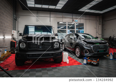 A black Mercedes G Class with hood open and a gray Audi SUV parked on a red and black checkered floor in a clean, well lit car shop garage. A black Mercedes G Class with hood open and a gray Audi SUV parked on a red and black checkered floor in a clean, well lit car shop garage. 134284992