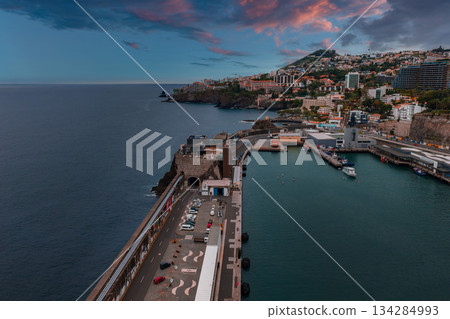 Wide angle view of Funchal harbor, Madeira, Portugal at dusk, with marina boats, fort like stone gateway, terraced houses, hotels, pink clouds, and calm teal water. Wide angle view of Funchal harbor, Madeira, Portugal at dusk, with marina boats, fort like stone gateway, terraced houses, hotels, pink clouds, and calm teal water. 134284993