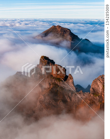Jagged volcanic peaks rise above thick clouds in Madeira, warm sunrise light on rust red rock, cool blue mist recedes, layered depth from elevated vantage 134285009