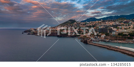 Panoramic coastal view of Funchal, Madeira, Portugal, with harbor, breakwater, marina, volcanic headlands, and white terracotta buildings rising on green hills. 134285015