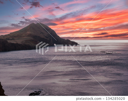 Rugged headland extends into the Atlantic at Ponta de Sao Lourenco, Madeira. A white structure rests below dark slopes, rocky islets dot calm water, and low light warms clouds. 134285020