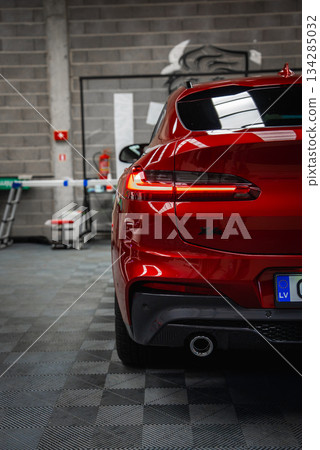 Shiny red BMW X4 parked in a well lit garage with tiled floor, gray brick wall, fire extinguisher, and visible European license plate marked LV. Shiny red BMW X4 parked in a well lit garage with tiled floor, gray brick wall, fire extinguisher, and visible European license plate marked LV. 134285032