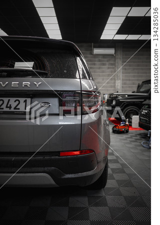 Silver Land Rover Discovery parked indoors with visible license plate, grid ceiling, tiled floor, and luxury cars in an active workshop setting. Silver Land Rover Discovery parked indoors with visible license plate, grid ceiling, tiled floor, and luxury cars in an active workshop setting. 134285036