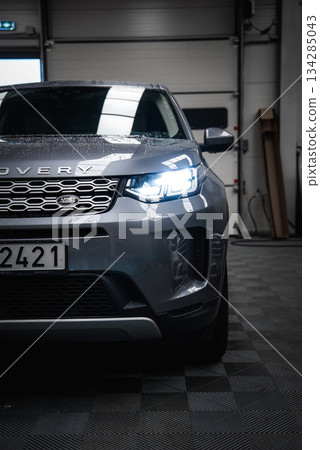 Silver Range Rover Discovery with illuminated headlights parked on a checkered floor inside a garage. Wet surface suggests recent cleaning or rain exposure. Silver Range Rover Discovery with illuminated headlights parked on a checkered floor inside a garage. Wet surface suggests recent cleaning or rain exposure. 134285043
