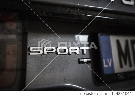 Detailed view of a luxury car's rear, showcasing the SPORT badge, part of the LV license plate, and reflective metallic paint under subtle lighting. Detailed view of a luxury car's rear, showcasing the SPORT badge, part of the LV license plate, and reflective metallic paint under subtle lighting. 134285044