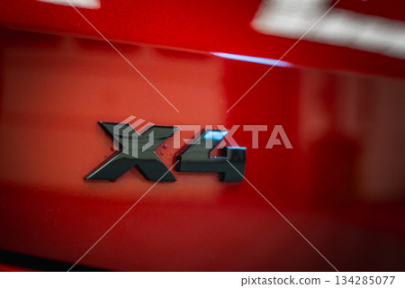 A detailed view of the black X4 badge on the rear of a red BMW. The glossy paint reflects light, with a blurred indoor showroom background. A detailed view of the black X4 badge on the rear of a red BMW. The glossy paint reflects light, with a blurred indoor showroom background. 134285077