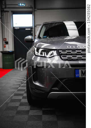 Silver Range Rover Discovery with water droplets on the hood, parked in a garage with a black door, green trash bin, and red carpet strip visible. 134285082