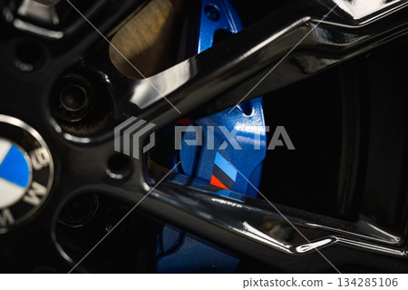 Detailed view of a BMW M series wheel featuring a glossy black alloy rim and blue brake caliper with the iconic tri color M logo in a showroom setting. 134285106