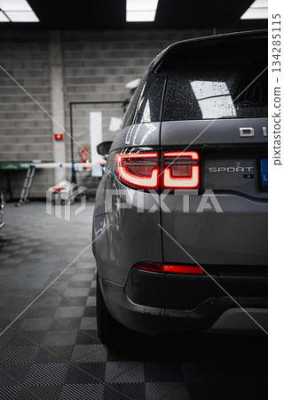 Gray Range Rover Discovery Sport with illuminated taillights in a well lit garage. Water droplets on the surface and tools visible in the background. Gray Range Rover Discovery Sport with illuminated taillights in a well lit garage. Water droplets on the surface and tools visible in the background. 134285115