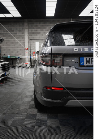 Silver Range Rover Discovery Sport parked in a modern garage with tiled floor, concrete walls, tools, and bright ceiling lights reflecting on the car. 134285124
