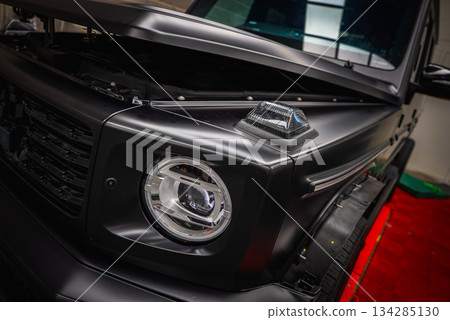 Front section of a black Mercedes G Class SUV indoors, featuring circular LED headlight, turn signal indicator, and matte finish on red carpet flooring. Front section of a black Mercedes G Class SUV indoors, featuring circular LED headlight, turn signal indicator, and matte finish on red carpet flooring. 134285130