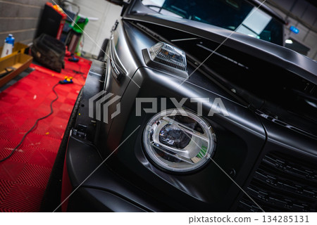 Front section of a black Mercedes G Class SUV with hood open, parked on a red checkered floor. Tools, a green hose, and supplies are visible in the garage. Front section of a black Mercedes G Class SUV with hood open, parked on a red checkered floor. Tools, a green hose, and supplies are visible in the garage. 134285131