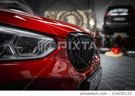 The front end of a red BMW X4 is displayed, highlighting its headlight and kidney grille. A checkered floor, tools, and a vehicle on a lift are visible. 134285132