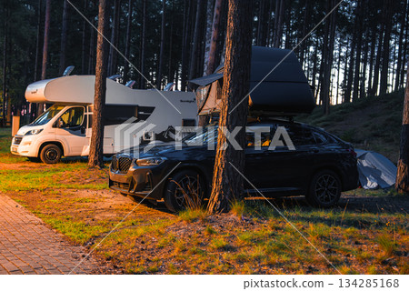 A black BMW X4 2025 with a rooftop tent parked on grass near a paved path, surrounded by tall pine trees and a white camper van at sunrise or sunset. A black BMW X4 2025 with a rooftop tent parked on grass near a paved path, surrounded by tall pine trees and a white camper van at sunrise or sunset. 134285168