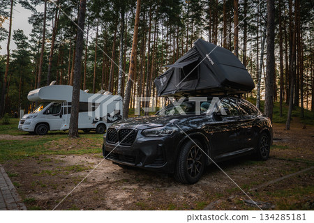 A black BMW X4 2025 with a rooftop tent is parked in a pine forest camping area. A white camper van and soft sunlight enhance the natural setting. 134285181