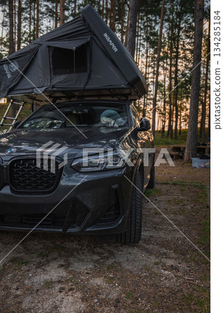 A BMW X4 2025 with a Kampa rooftop tent parked in a forested camping area. Tall pine trees, a picnic table, and warm sunset lighting are visible. 134285184