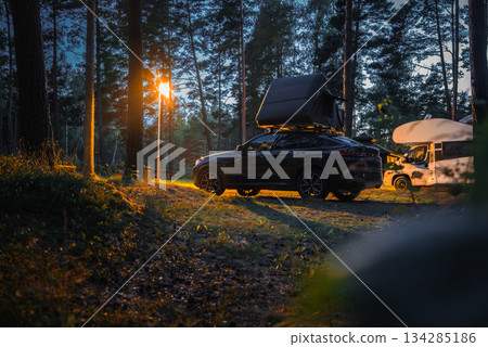 A BMW X4 2025 with a rooftop tent parked in a twilight forest camping area. A warm streetlight illuminates the scene, with trees and a camper van nearby. A BMW X4 2025 with a rooftop tent parked in a twilight forest camping area. A warm streetlight illuminates the scene, with trees and a camper van nearby. 134285186