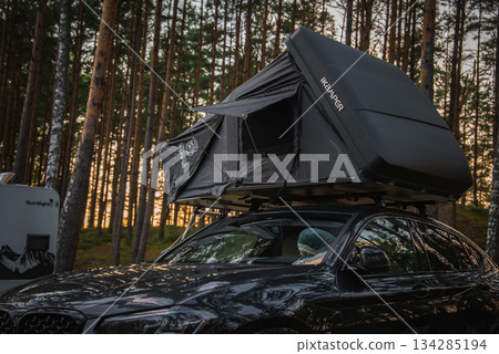 A black BMW X4 2025 with an IKAMPER rooftop tent parked in a forested area. Tall pine trees and a camper van are visible during a warm sunset. 134285194