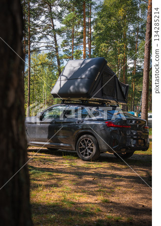 A BMW X4 2025 parked in a forest surrounded by tall pine trees, with a rooftop tent partially open under sunlight filtering through the branches. 134285214