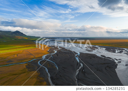 Braided river channels flow through dark sediment in Iceland's open landscape, with a green hillside and golden sunset hues under a cloudy sky. Braided river channels flow through dark sediment in Iceland's open landscape, with a green hillside and golden sunset hues under a cloudy sky. 134285231