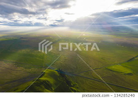 Aerial view of a green Icelandic landscape at sunset, featuring a grassy hill, scattered clouds, sunlight beams, a straight road, and small water bodies. Aerial view of a green Icelandic landscape at sunset, featuring a grassy hill, scattered clouds, sunlight beams, a straight road, and small water bodies. 134285240