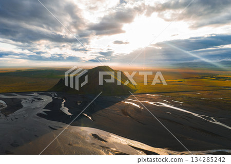 A solitary hill rises from a flat landscape in Iceland, surrounded by winding black sand riverbeds. Sunset light highlights green and earthy tones. 134285242