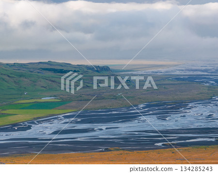 Aerial view of Iceland's braided river system with volcanic sands, green farmland, rugged cliffs, and a coastline under low hanging clouds at sunset. Aerial view of Iceland's braided river system with volcanic sands, green farmland, rugged cliffs, and a coastline under low hanging clouds at sunset. 134285243