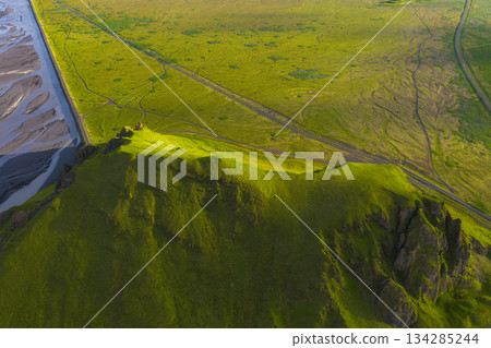 Aerial view of a green Icelandic landscape with a grassy hill, rugged rocks, and a braided river system illuminated by warm sunset light. Aerial view of a green Icelandic landscape with a grassy hill, rugged rocks, and a braided river system illuminated by warm sunset light. 134285244