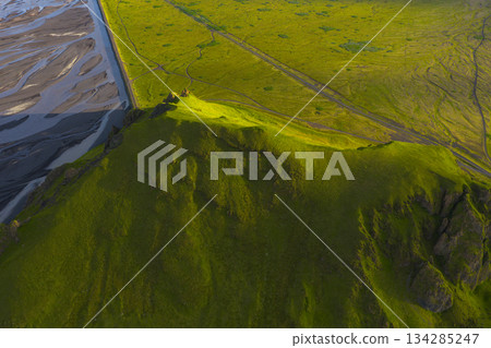 Aerial view of a grassy hill with rugged slopes, a braided river system, and flat terrain, illuminated by warm sunset light in Iceland. 134285247