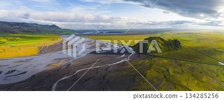 Aerial view of a green hill rising from flat terrain, a braided river system, vibrant fields, volcanic soil, and a road under a partly cloudy sunset sky. 134285256