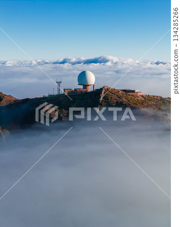 Aerial view shows a white geodesic radar dome and masts on rocky ridges at Pico do Arieiro, Madeira, Portugal. Soft low sun light casts warm tones over clouds below. Aerial view shows a white geodesic radar dome and masts on rocky ridges at Pico do Arieiro, Madeira, Portugal. Soft low sun light casts warm tones over clouds below. 134285266