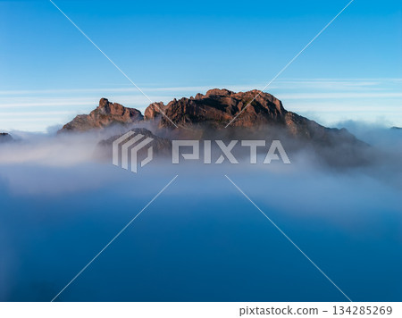 Rugged reddish cliffs rise above a cloud layer at dawn in Pico do Arieiro, Madeira, Portugal. Soft light reveals sharp ridges and texture, with a clear blue sky. 134285269