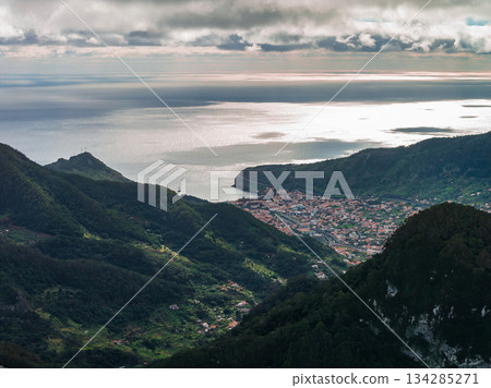 High viewpoint looks over green mountains to a bay and compact town of Sao Vicente, Madeira, Portugal. Low clouds part and light casts silver on Atlantic and slopes. High viewpoint looks over green mountains to a bay and compact town of Sao Vicente, Madeira, Portugal. Low clouds part and light casts silver on Atlantic and slopes. 134285271