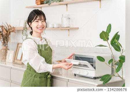 一位年輕的亞裔女性，穿著圍裙，正在廚房使用烤麵包機。 134286676