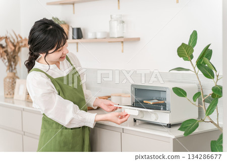 一位年輕的亞裔女性，穿著圍裙，正在廚房使用烤麵包機。 134286677