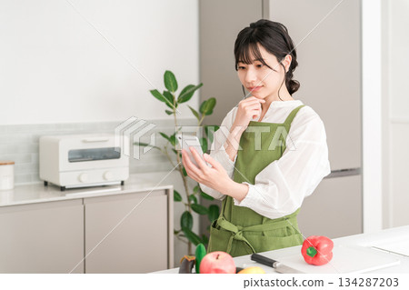 A young Asian woman wearing an apron cooking in the kitchen while looking at her smartphone A young Asian woman wearing an apron cooking in the kitchen while looking at her smartphone 134287203