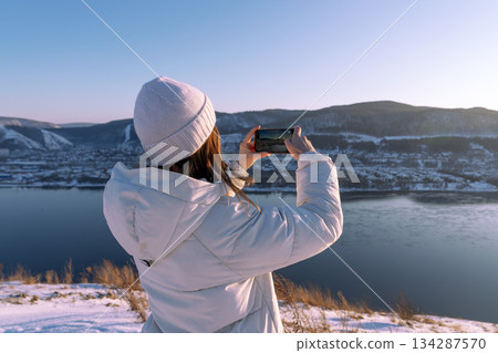 A woman skillfully captures the stunning beauty of a winter landscape using her smartphone by a tranquil river. Photographing nature on a winter adventure trip - A girl in a white jacket takes A woman skillfully captures the stunning beauty of a winter landscape using her smartphone by a tranquil river. Photographing nature on a winter adventure trip - A girl in a white jacket takes 134287570