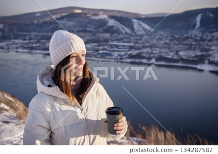 A woman enjoys sipping warm coffee, completely surrounded by captivating winter landscapes and a peaceful river view, A woman in a white jacket and hat drinks hot coffee in winter by the riverbank and A woman enjoys sipping warm coffee, completely surrounded by captivating winter landscapes and a peaceful river view, A woman in a white jacket and hat drinks hot coffee in winter by the riverbank and 134287582