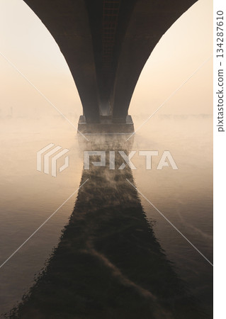 The arch of the bridge is reflected in the water, an architectural element of art. Old arched bridge over the Yenisei River, which does not freeze in winter, against the background of a foggy sunset 134287610
