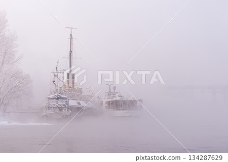 Atmospheric foggy landscape with nostalgic mood. Old decommissioned vessels, a Jung river tram and a tugboat or a river icebreaker permanently parked on the Yenisei River, shrouded in mystery and fog 134287629
