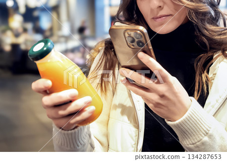 A young blonde white woman buys juice in a modern supermarket. She reads the label with her phone, scans barcodes and examines the ingredients. Healthy eating and healthy lifestyle. 134287653