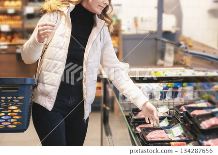 Blonde woman in a white jacket happily picks out red meat from the counter at a well-organized supermarket. She carries a shopping basket, enjoying her grocery shopping experience 134287656