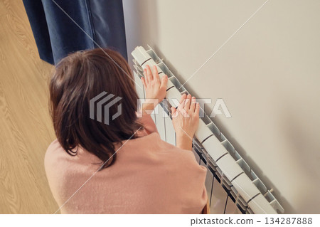 Woman feeling cold and touching white radiator for warmth inside home 134287888