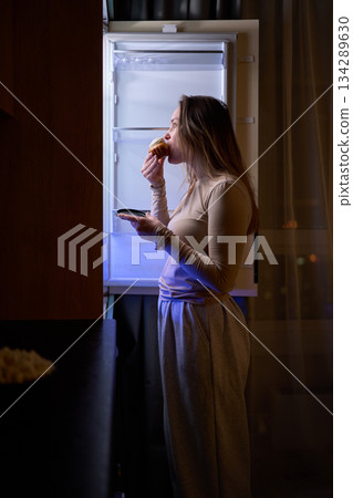 Female eating muffin while standing in front of open refrigerator Female eating muffin while standing in front of open refrigerator 134289630