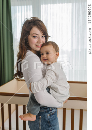 Cozy family moment with a young woman holding her smiling baby at home, standing near soft window light. A warm and cozy parent-child scene filled with love 134290408